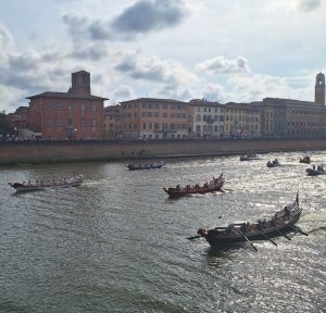 Regata delle Repubbliche Marinare sul fiume Arno a Pisa. In primo piano, il galeone verde di Venezia è in testa alla gara, seguito dai galeoni di Genova (bianco), Pisa (rosso) e Amalfi (blu). Le sponde del fiume sono gremite di spettatori davanti ai palazzi storici.